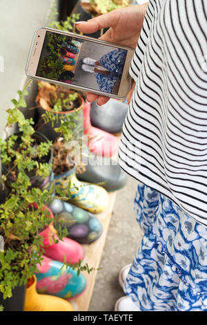 Nahaufnahme von einem Mädchen, das Aufnehmen von Bildern auf einem Smartphone bunte Gummistiefel als Blumentöpfe mit verschiedenen blühenden Blumen auf der Veranda, Konzept der Stockfoto