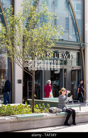 Eine attraktive junge Frau, Sitzen, trinken ein Costa takeaway Kaffee außerhalb John Lewis Store in Cheltenham UK beim Prüfen der Handy. Stockfoto