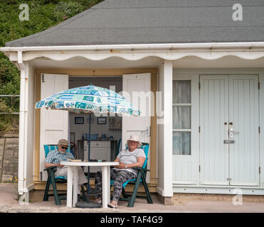 Älteres Rentnerehepaar sitzen auf Liegen am Strand liegen ausserhalb ihrer Beach Hut in Bournemouth nahe der Mitte Chine und Alum Chine unter dem Dach Stockfoto