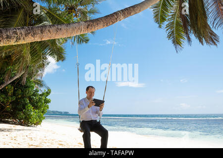 Mann sitzt auf Schaukel mit digitalen Tablet am Sandstrand in der Nähe der idyllischen Meer Stockfoto