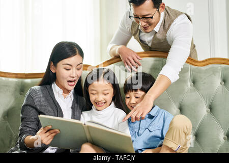 Junge asiatische Eltern und zwei Kindern auf der Couch lesen Buch sitzen zusammen in der Familie Wohnzimmer zu Hause Stockfoto