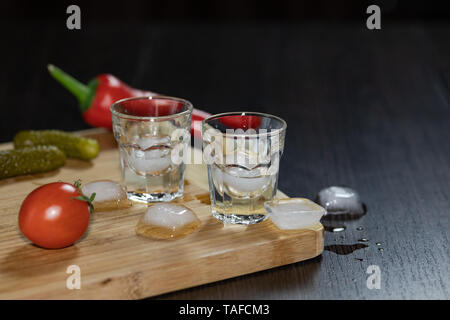 Zwei Shot Gläser Wodka auf dem Schneidbrett mit Kirschtomaten, marinierten Gurken, Paprika und Eiswürfel. Schwarzer Hintergrund Stockfoto