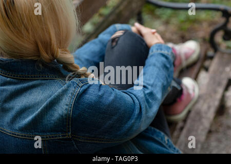 Blonde Frau in Jeans Jacke sitzt allein auf einer Parkbank, hält ihr Bein mit ihren Armen Stockfoto