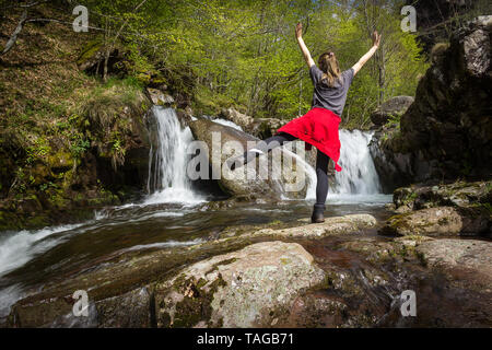 Mädchen mit roten Jacke um ihre Taille Stehen auf einem Bein mit den Händen in der Luft vor der malerischen Wasserfall an einem Bergbach im frühen Frühjahr Stockfoto