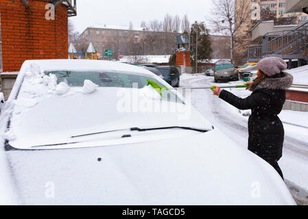 Junge schöne Frau in einem schwarzen Mantel reinigt den Schnee mit einem klappbaren Grüne lange Bürste von der Oberfläche von ihrem Auto Stockfoto