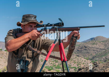 Ein professioneller Jäger zielt darauf ab, eine Schrotflinte Gewehr mit einem Stativ auf einer Farm in Namibia unterstützt. Stockfoto