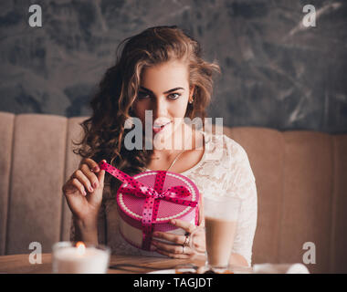 Lächelnd schöne Frau öffnen vorhanden Box mit Pink Ribbon im Zimmer sitzen. Mit Blick auf die Kamera. Valentines Tag. Stockfoto