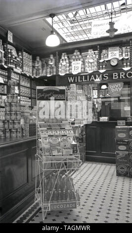 1950er, historisch, Innenansicht eines Williams Brothers Grocery and Proviures Stores, England, Großbritannien, mit Produkten der Epoche, darunter Obst aus der Dose, Kürbis, Puderzucker, hoch gestapelte Erdbeermarmelade und ein Schild für „Divi Day“ am nächsten Dienstag. Außerdem gibt es einen Ausstellungsstand für Lucozade, das Getränk mit Kohlensäure, das zur Heilung von Krankheiten beworben wurde. Traditionelle Lebensmittelläden für Familien und Versorgungsgeschäfte wie dieses waren ein typisches Fest des britischen Hochhauses in dieser Zeit, das erst Ende der 1960er Jahre aufgrund der Konkurrenz aus den aufstrebenden Supermärkten zurückging. Stockfoto