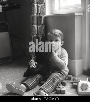 1960, historische, ein Junge, ein Paar "Rupert Bär" Hosen und Hausschuhe in der Ecke eines Zimmers sein Kuscheltier Hund umarmt sitzen und neben ihm sein Spielzeug Ziegel und Telefon, England, UK. Stockfoto