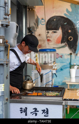 Junger Mann Anbieter kochen Pfannkuchen Buchimgae oder Koreanisch, Koreanisch traditionelles Essen, an einer Garküche in Gamcheon Culture Village in Busan, Südkorea Stockfoto