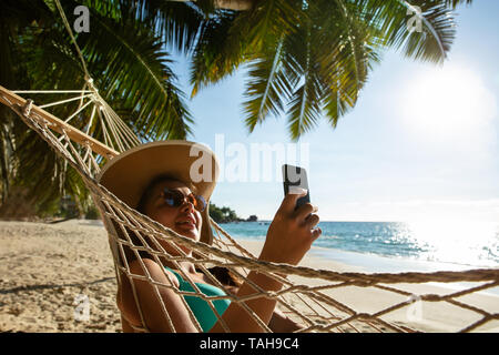Lächelnden jungen Frau mit Hut entspannen auf der Hängematte mit Handy am Strand Stockfoto