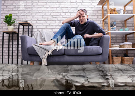 Verärgert junge Mann sitzt auf einem Sofa im Wohnzimmer überschwemmt mit Wasser Stockfoto