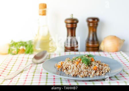 Buchweizen Brei mit Karotten und Dill in grau Platte. Auf dem Hintergrund der Goldenen zwiebeln, grüne Petersilie und Salz und Pfeffer Mühlen. Stockfoto