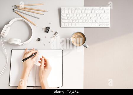 Hände von Frau schreiben im Notebook, PC-Tastatur, Kopfhörer und Tasse Kaffee auf grauem Hintergrund, Ansicht von oben Stockfoto