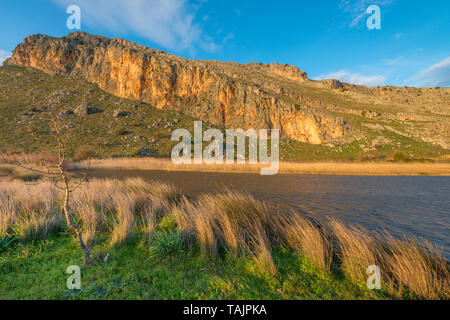 Sonnenuntergang in Griechenland, goldenen Stunde in die Berge, kleiner See mit geblasenem Gras, starker Wind. Schiere Berge wand mit goldfarbenen Rock. Stockfoto