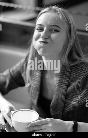 Spaß junge blonde Frau mit Tasse Kaffee im Café mit Blick auf die Kamera lächelnd, grimassen Schwarzweiß Stockfoto