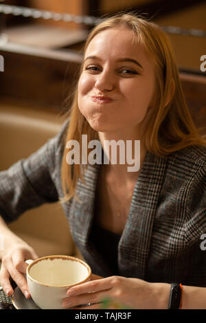 Spaß junge blonde Frau mit Tasse Kaffee im Café mit Blick auf die Kamera lächelnd, Grimassen Stockfoto