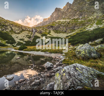 Bergsee mit Wasserfall und Felsen im Vordergrund bei Sonnenuntergang. Velicka Tal, Hohe Tatra, Slowakei. Stockfoto