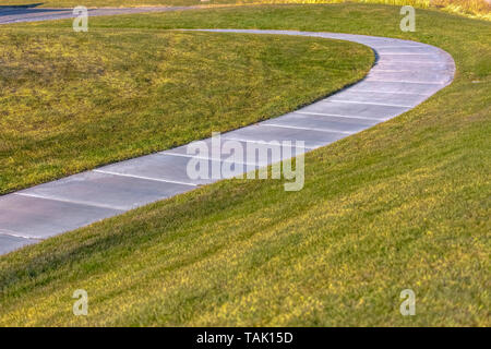 Gepflasterter Fußweg, die Kurven durch die grasbewachsenen Gelände auf einem sonnigen Tag gesehen. Der Weg führt zu einer Straße, die durch helles Sonnenlicht beleuchtet wird. Stockfoto