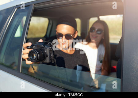 Blogger in Gläsern seiner Reise vlog Nehmen auf professionelle Kamera auf Road Trip. Mann Fotograf die Bilder aus offenen Fenster mit Freundin Stockfoto