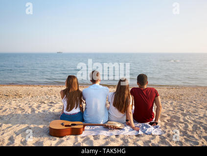Zurück Blick auf eine Gruppe von Freunden zusammen sitzen auf dem Handtuch auf weißen Sandstrand im Urlaub und genießen den Sonnenuntergang über dem Meer. Männer und Frauen Stockfoto