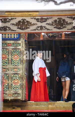 Miko, oder shrine Maiden, steht am Eingang zum Toshogu Schrein der Gebetshalle (Haiden), mit Touristen, an einem regnerischen Tag in Nikko, Tochigi, Japan. Stockfoto