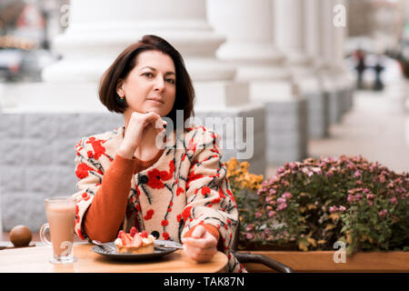 Stilvolle Frau 40-45 Jahre alten Kuchen essen und trinken Kaffee im Cafe im Freien. Mit Blick auf die Kamera. Stockfoto