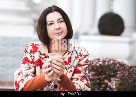 Lächelnd reife Frau 45-50 Jahre alten Holding Tasse Kaffee sitzen im Cafe im Freien. Mit Blick auf die Kamera. Frühling Saison. Stockfoto