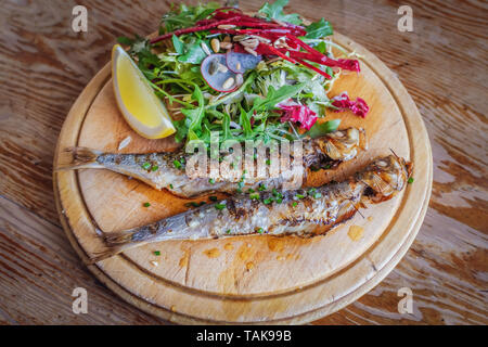 Ganzen gegrillten Sardinen auf einer hölzernen Platte mit einem frischen Salat und Zitrone in Scheiben schneiden, auf einem Holztisch mit einem attraktiven Korn serviert. Stockfoto