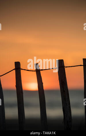 Die untergehende Sonne, die durch einen Zaun auf einem sandigen Strand gesehen Stockfoto