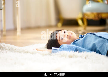 Süße kleine asiatische Junge lag auf dem Rücken auf dem Teppich im Wohnzimmer sah lächelnd