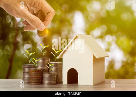 Eigenschaft Investition oder Geld sparen für zu Hause kaufen. Ein Geschäftsmann Hand Münze über wachsende Pflanze auf Gestapelte Münzen und Resident house Modell auf Holz Stockfoto