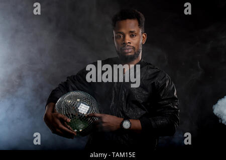 Headshot von schweren Dunkelhäutige männliche Holding Kinn geheimnisvoll Kamera suchen, in schwarz windjacke Jacke, wristwatces gekleidet, gegen die ständigen Stockfoto