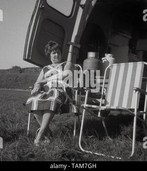 1960, historische, oh die einfachen Freuden des Lebens.... eine Dame entspannten Sitzen in einem Metall Klappstuhl an der Rückseite eines Morris Commerical J4 Van in einem Feld mit der Rückseite mit offenen Türen, England, UK geparkt Stockfoto