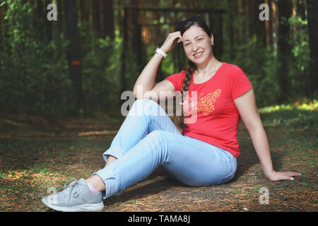 Junge schöne Frau sitzt auf dem Boden und das Gefühl glücklich. Sie ist legere Kleidung. Wald Hintergrund. Lebensstil Stockfoto