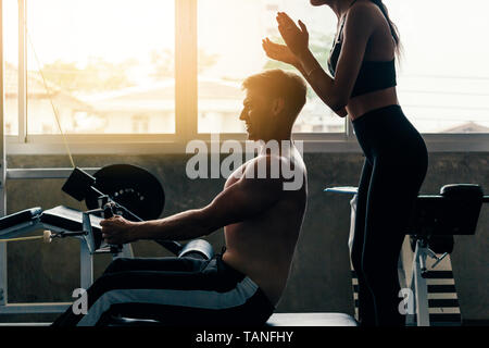 Jungen kaukasischen Mann im Hemd an einer Turnhalle, hartem Training und Gewichten in eingesetztes Reihe Maschine ziehen mit Unterstützung von weiblichen Fitness personal trainer Stockfoto