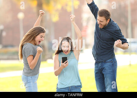 Vorderansicht Portrait von drei aufgeregt Freunde springen Kontrolle smart phone in einem Park Stockfoto