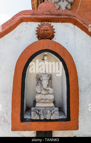 Dusun Ambengan, Bali, Indonesien - 25. Februar 2019: Kleine graue Stein Lord Ganesha Statue, die in roten Rahmen Nische in der weißen Wand gesetzt. Lächelnde Sonne Bild auf Stockfoto