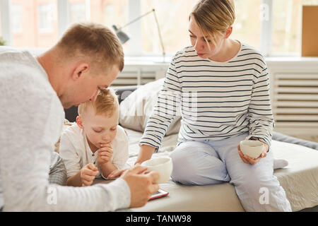 Acht junge Eltern in homewear saß auf dem Bett und Unterstützung der Sohn mit lernspiel auf Tablet beim Trinken von Kaffee am Morgen Stockfoto