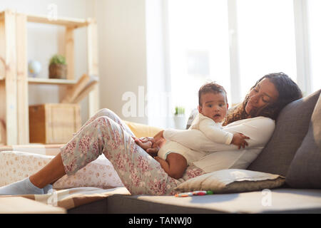 Lächelnd sorgfältige liebevolle junge afrikanische Mutter mit gewellten Haar sitzen auf komfortablen Sofa und umarmen Baby, während Sie gemeinsam entspannende Stockfoto