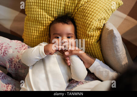 Oben Ansicht von adorable African baby boy in niedliche Pullover liegen auf Mütter Beine und beißt sich in den Finger, weil der Dentition Stockfoto