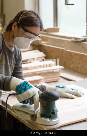 Junge Frau schleift das Holzbrett mit kantigen Schleifmaschine in der Werkstatt Stockfoto