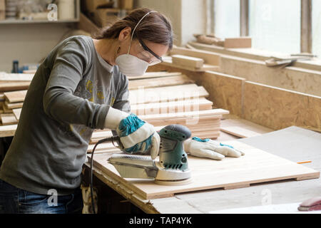 Junge Frau schleift das Holzbrett mit kantigen Schleifmaschine in der Werkstatt Stockfoto