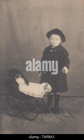 Vintage Fotostudio Postkarte zeigt ein junges Mädchen mit ihrem Spielzeug Puppe in Ihr Spielzeug Kinderwagen Stockfoto