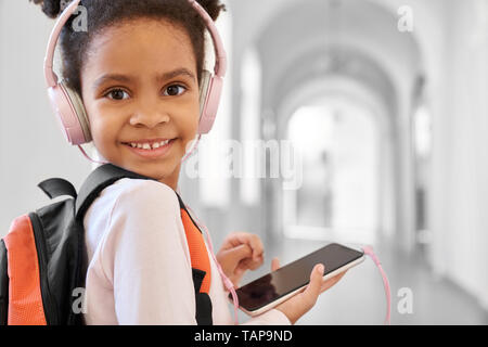 Schön, positive Schulmädchen mit Schule Rucksack und Kopfhörer holding Telefon und lächelnd. Glücklich, schönes Kind stehen in der Halle der Grundschule, an der Kamera suchen, posieren. Stockfoto