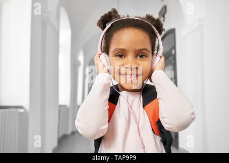 Positive, schöne, freundliche Schulmädchen mit Kopfhörern auf dem Flur der Schule prrimary posieren. Hübsches, lächelnde Mädchen hören Musik in der Pause zwischen den Unterrichtsstunden. Stockfoto