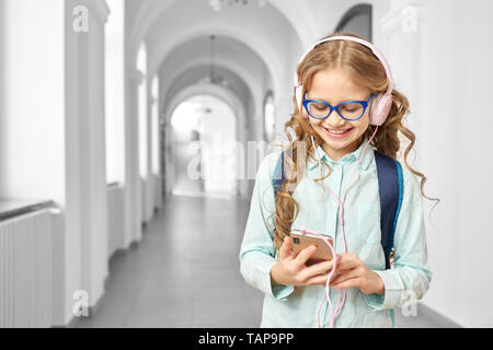 Schön, schöne Schule Mädchen Musik hören mit Kopfhörern in der Pause zwischen den Unterrichtsstunden. Happy girl in Gläsern, Telefon, lächelnd und stand auf dem Flur der Schule. Stockfoto
