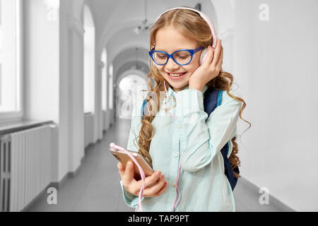 Fröhliches Mädchen in Blau Brille mit Rucksack stand auf dem Flur der Schule, Holding und Telefon. Ziemlich schulkind Musik hören mit Kopfhörern mit Vergnügen.