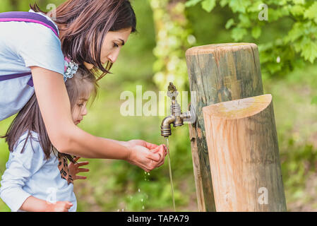 Junge Mama hilft ihr kleines Getränke Wasser aus einem Holz im Wald tippen. glückliche Mutter und Tochter Momente mit Liebe und natürliches Gefühl Stockfoto