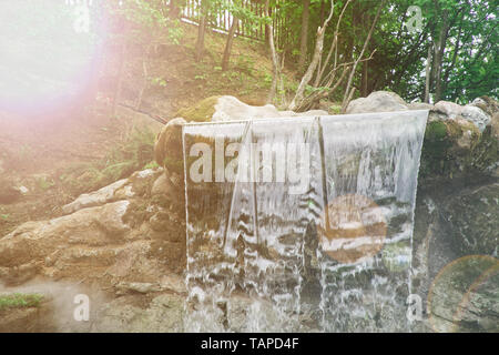 Garten Wasserfall von Steinen. Das Wasser fließt durch den Stein. Die Morgensonne scheint auf den Wasserfall Stockfoto